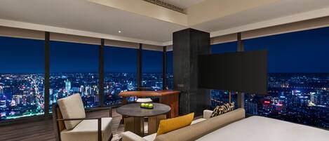 Une chambre d’hôtel moderne avec un grand lit, un canapé, une petite table avec des fruits et un téléviseur. La chambre offre une vue sur la ville grâce à de grandes fenêtres.