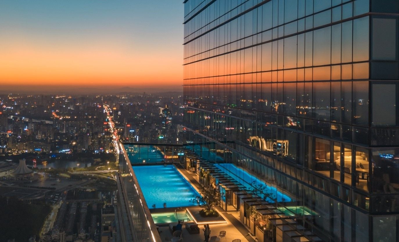 Un edificio alto con piscina en la azotea y zona de estar, rodeado por un paisaje urbano al atardecer.