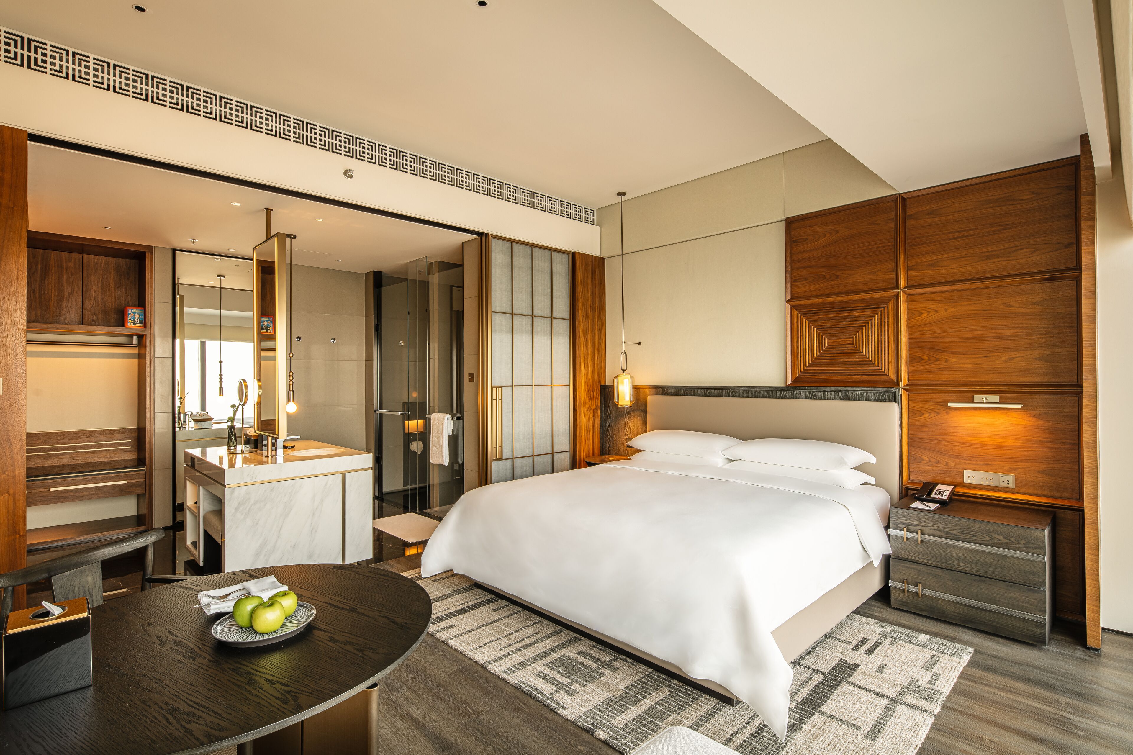 A modern hotel room with a large bed, a bedside table with a lamp, a mirror, a wooden wardrobe, and a bathroom with a glass shower.