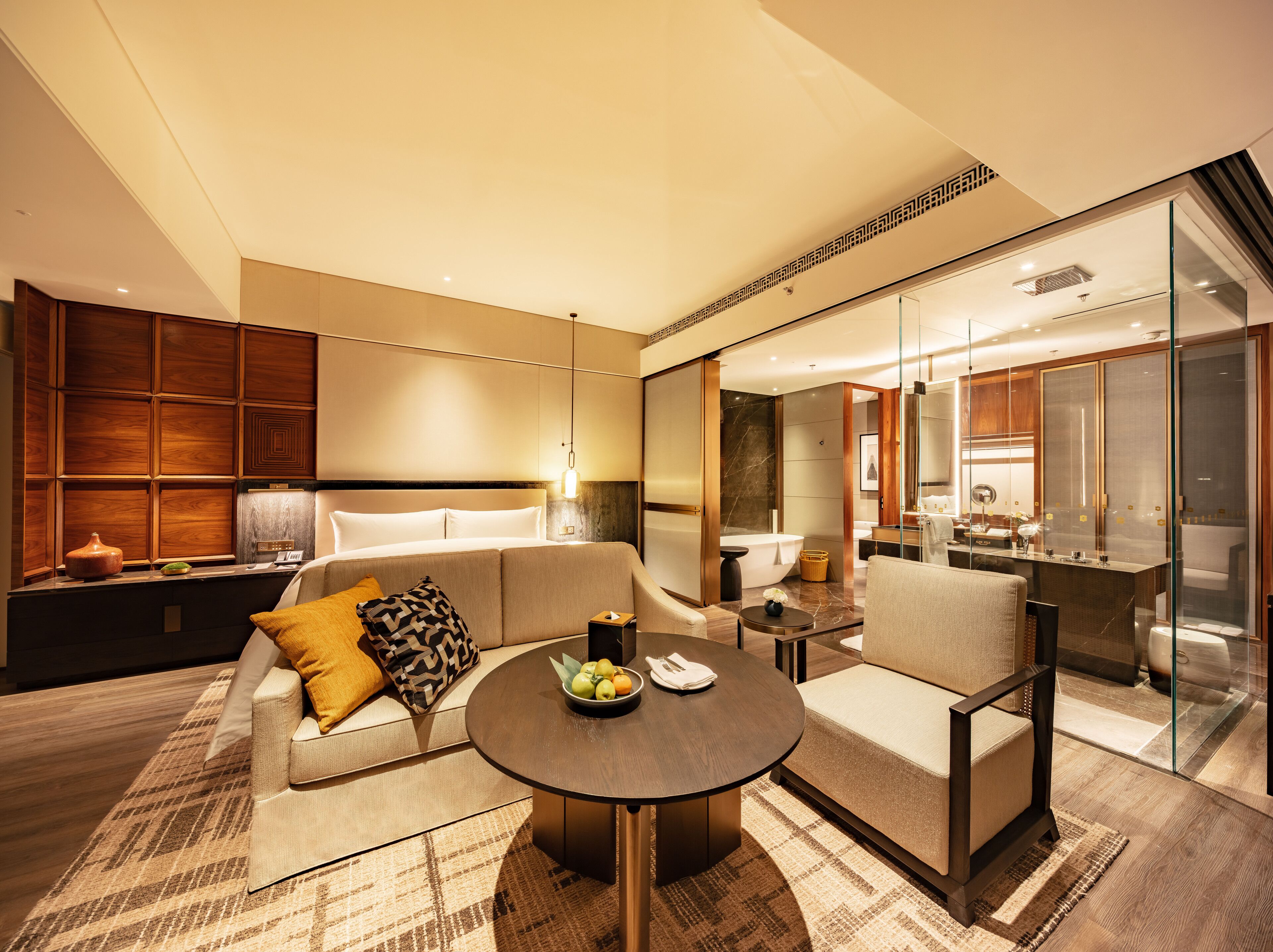 A modern hotel room with a sofa, armchairs, a round table, a small side table, and a glass-enclosed bathroom.