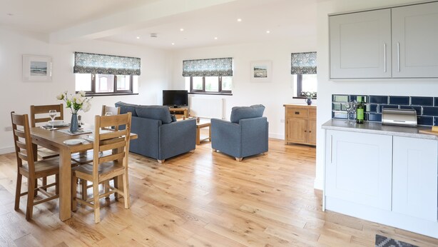 A spacious living area with a wooden dining table, blue sofas, and a kitchen with white cabinets. - Bwthyn Cae Haidd (Beaumaris)