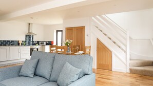 A modern living room with a grey sofa, a wooden dining table with chairs, a kitchen in the background, and a staircase leading to an upper floor. - Bwthyn Cae Haidd (Beaumaris)