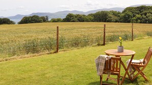 A grassy outdoor area with a table and chairs, a fence, and a view of a field and mountains. - Bwthyn Cae Haidd (Beaumaris)