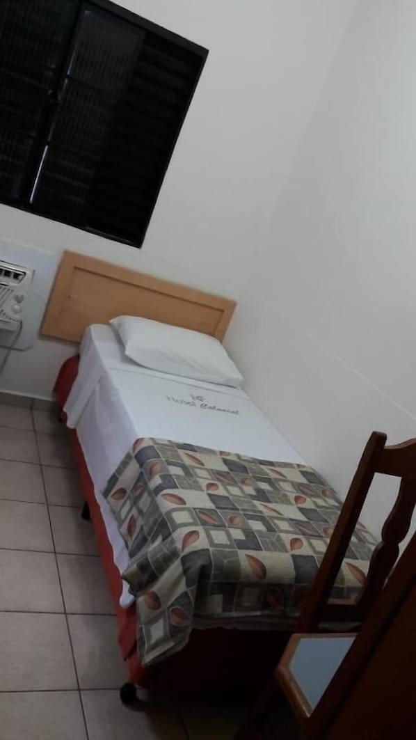 Una habitación pequeña con cama individual, cabecero de madera, colcha blanca y manta estampada. Se ve una ventana con persianas negras, una silla y una mesa pequeña con un mantel rojo.