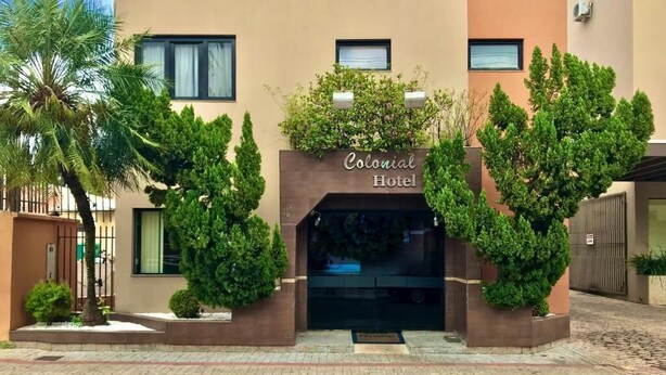 Fachada del Hotel Colonial con diseño moderno, que presenta una entrada central con un letrero, jardines simétricos y un camino de acceso con portón.