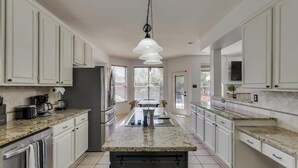 A spacious kitchen with white cabinets, granite countertops, a central island, and a hanging pendant light. - Spacious & Cozy Texas Gem with Private Pool (Plano)