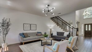 A spacious living room with a large sectional sofa, a coffee table, and two armchairs. A staircase with a black railing leads to an upper level. - Spacious & Cozy Texas Gem with Private Pool (Plano)