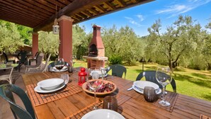 An outdoor dining area with a brick oven, wooden table, chairs, and a view of olive trees.