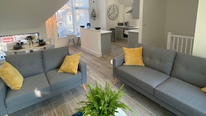 Uma sala de estar moderna com dois sofás cinzas, uma mesa de centro redonda com uma planta e um tapete azul estampado.