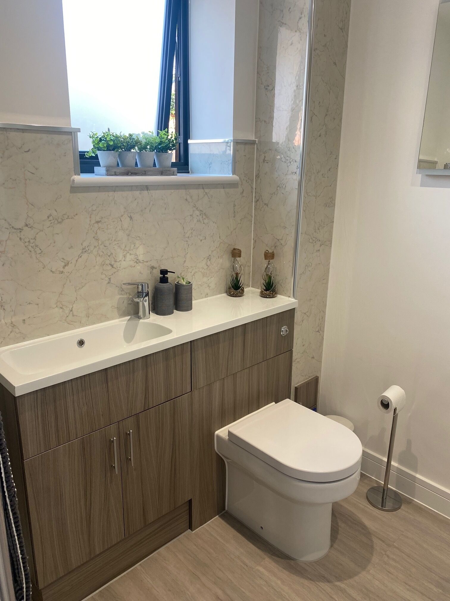 Ein modernes Badezimmer mit Marmorwaschbecken, Holzschränken, einer Toilette und einem Fenster mit Topfpflanzen.