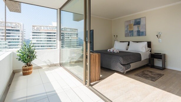 A modern bedroom with a bed, bedside tables, a small plant, and a view of the city through a balcony. - Arauco Wood - 2BR, Parking, Wifi, Mall & Pool (Vitacura)