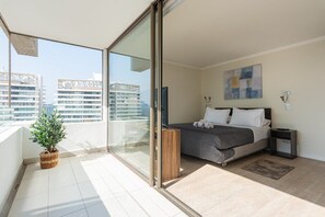Un dormitorio moderno con cama, mesitas de noche, una planta pequeña y vistas de la ciudad desde un balcón.