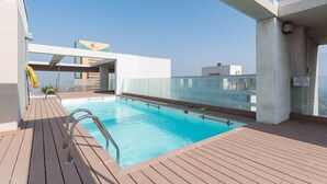 A rooftop pool with a wooden deck, clear skies, and a view of the cityscape. - Arauco Wood - 2BR, Parking, Wifi, Mall & Pool (Vitacura)