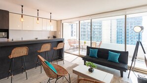 A modern kitchen with bar stools, a black sofa with blue cushions, and a large window offering a city view. - Arauco Wood - 2BR, Parking, Wifi, Mall & Pool (Vitacura)
