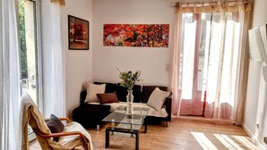 Een woonkamer met een bank, een glazen salontafel en een fauteuil. Aan de muur hangt een schilderij en er is een raam met gordijnen.