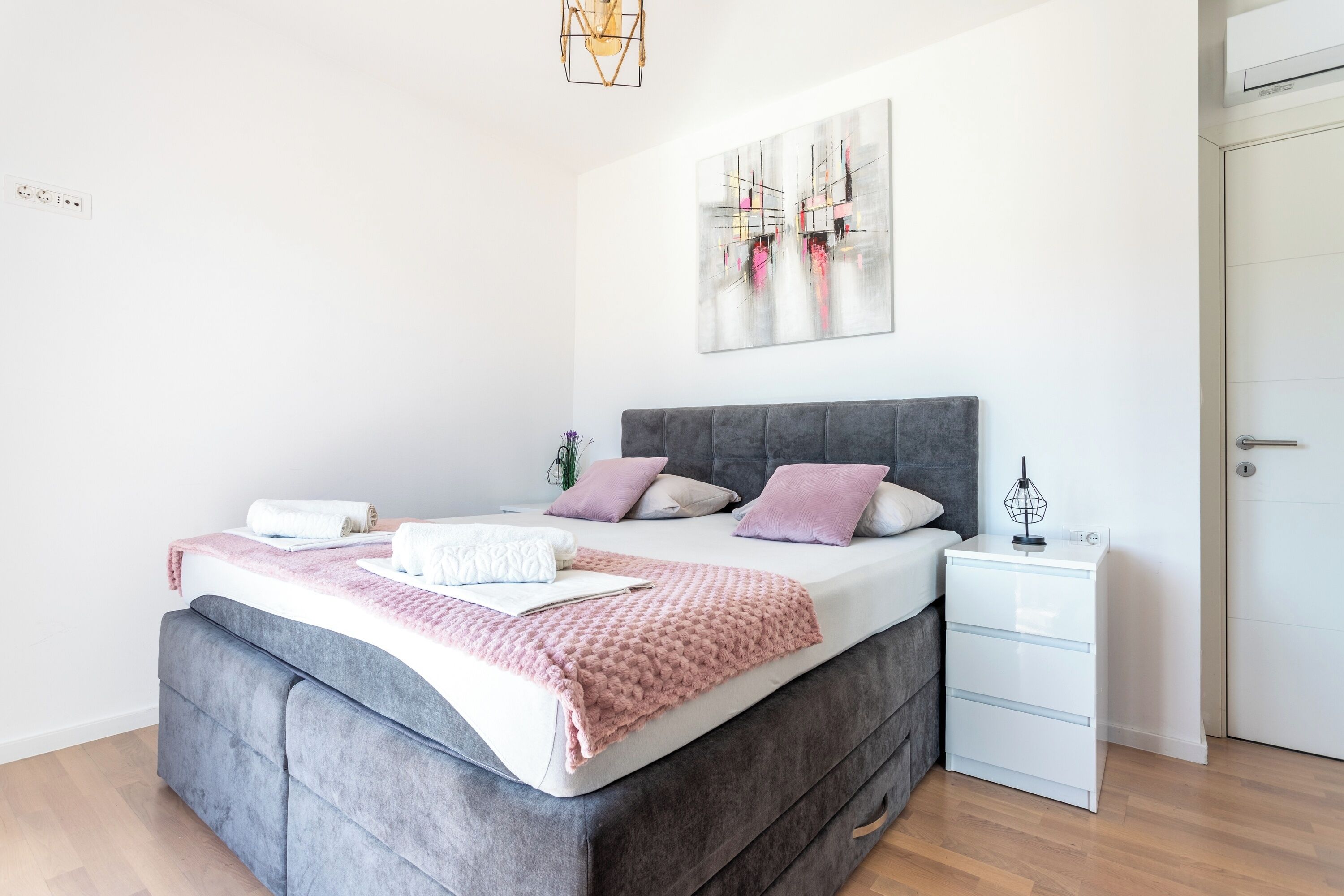 Une chambre à coucher moderne avec un lit recouvert d’un tissu gris, une table de chevet blanche et une tête de lit grise.