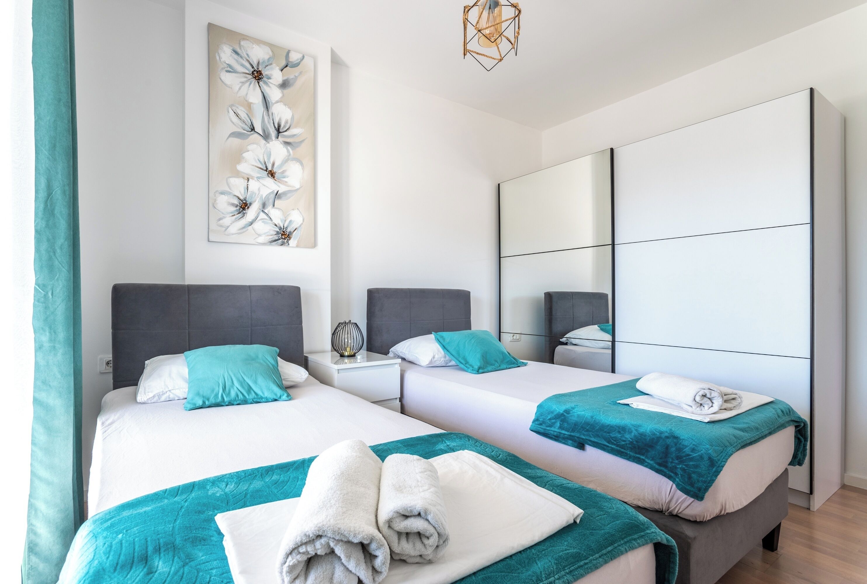 A modern hotel room with two beds, teal bedding, a large mirror, and a floral artwork on the wall.