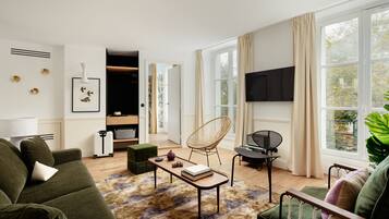 A modern living room with a sofa, a coffee table, a chair, and a TV. There is a window with curtains, a plant, and a door leading to another room.