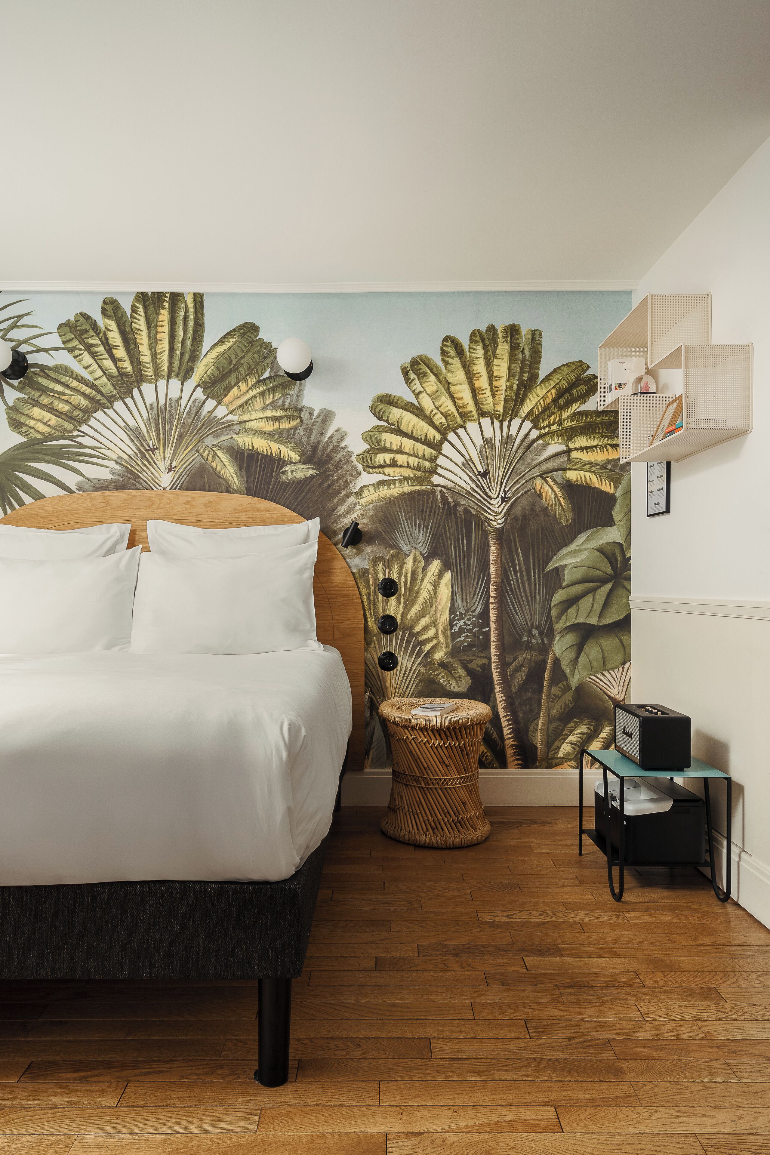 A bedroom with a bed, a wall mural, a small round table, a wicker chair, and a side table with a lamp.