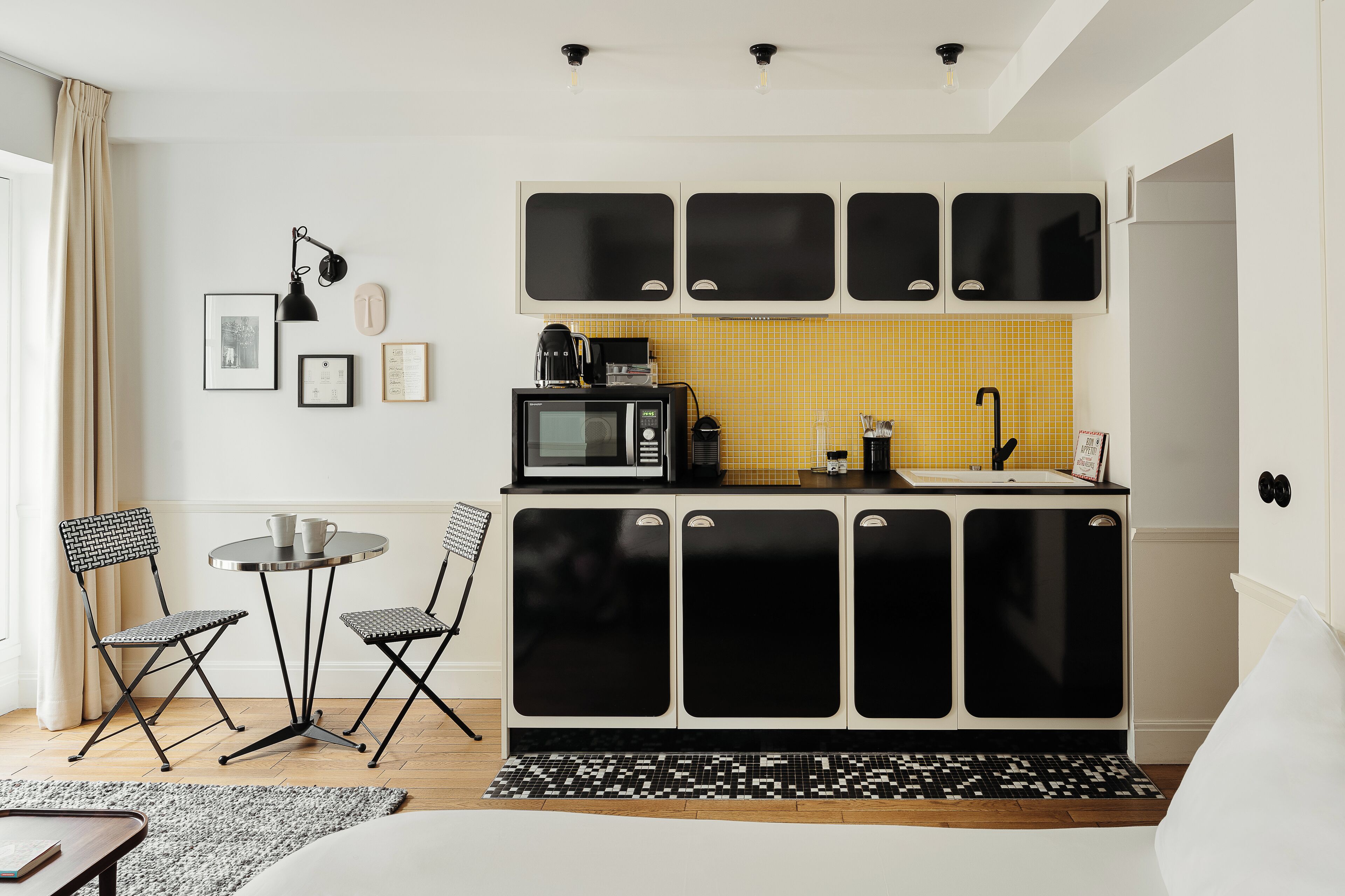 A compact kitchen with a microwave, coffee maker, and sink. There are two chairs and a small table. The kitchen has a yellow backsplash and black cabinets.