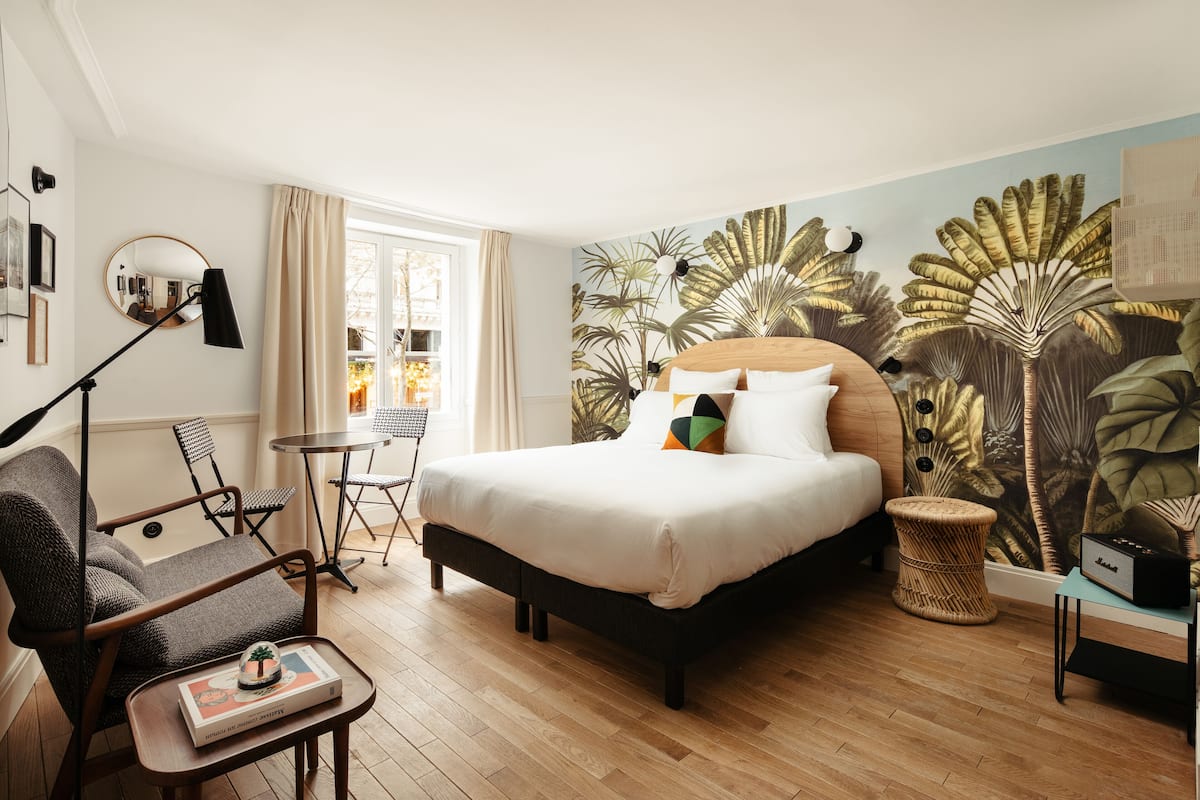 Ein Hotelzimmer mit einem großen Bett, einer Sitzecke mit Sessel und kleinem Tisch, einem runden Tisch und einer Wand mit einem tropischen Blatt-Wandgemälde.