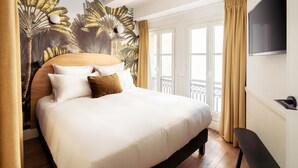 A hotel room with a large bed, a mural of tropical leaves on the wall, a TV mounted on the wall, and a balcony with curtains. - My Maison In Paris - Louvre (Paris)