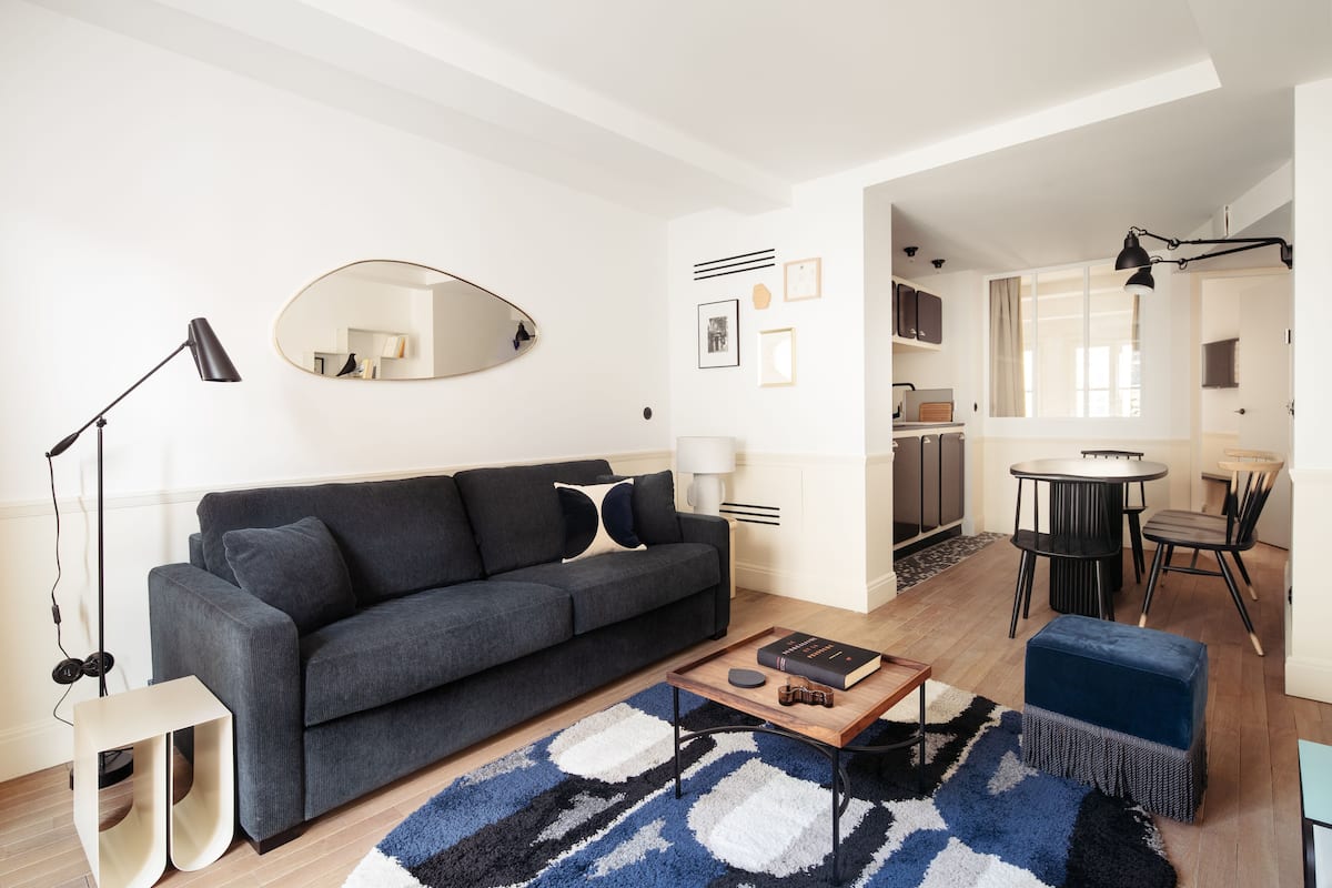 Ein modernes Wohnzimmer mit einem dunkelgrauen Sofa, einem Couchtisch und einem blauen Hocker. Im Hintergrund steht ein runder Tisch mit Stühlen, und an der Wand hängt ein Spiegel.