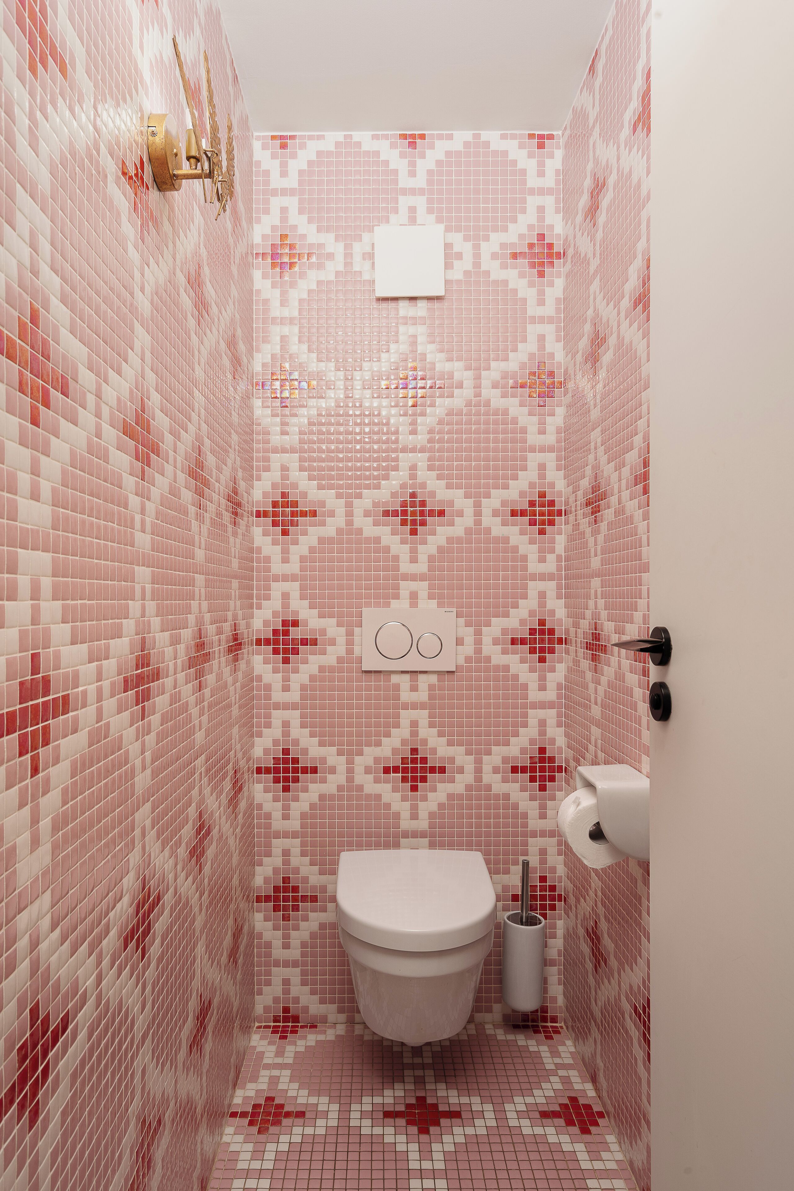 Une salle de bain avec des carreaux de mosaïque roses et blancs, des toilettes blanches, un distributeur d’essuie-tout et un luminaire fixé au mur.