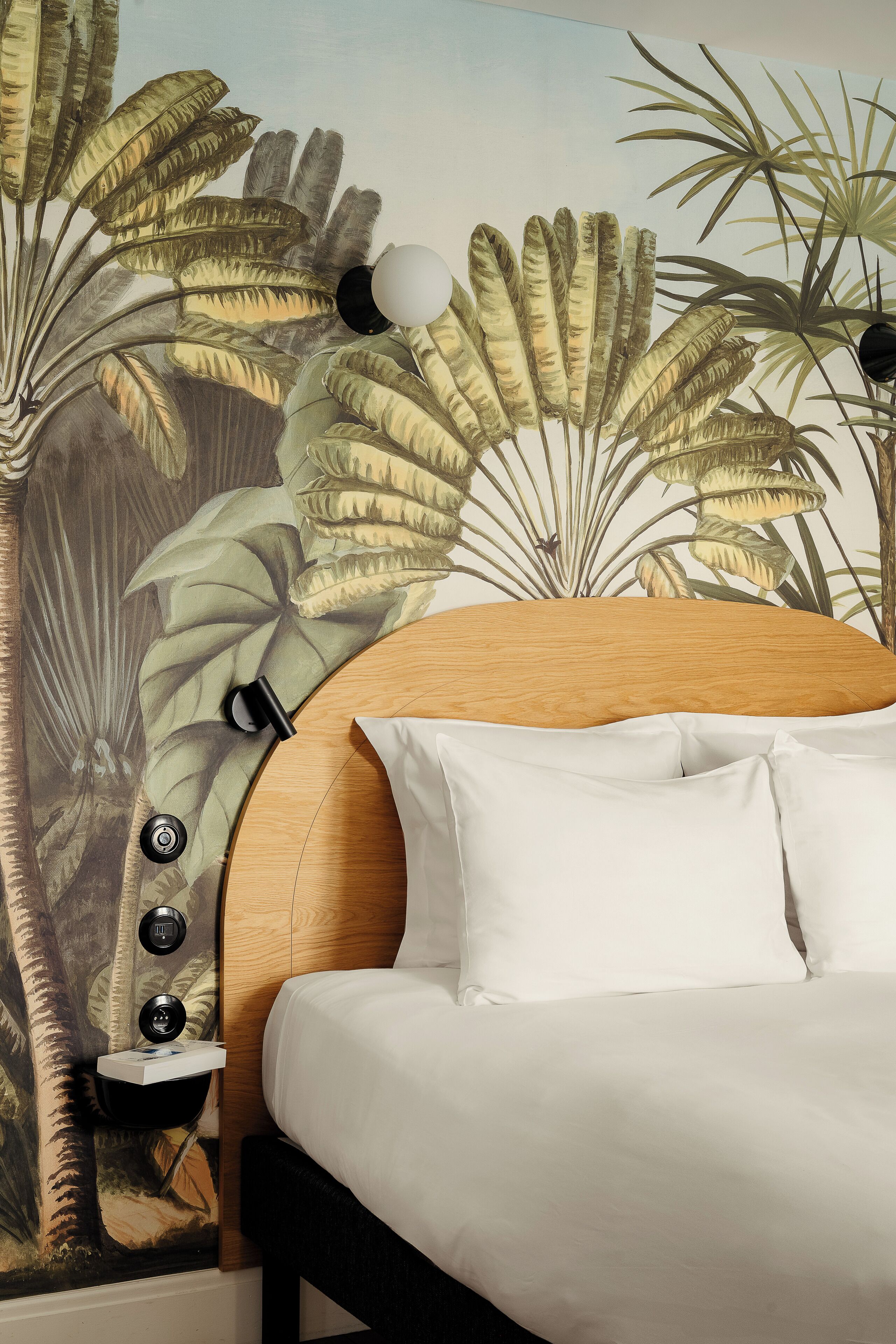 Une chambre avec un grand lit, du linge de lit blanc, une tête de lit en bois, une table de chevet avec une lampe et une fresque murale représentant une végétation tropicale.