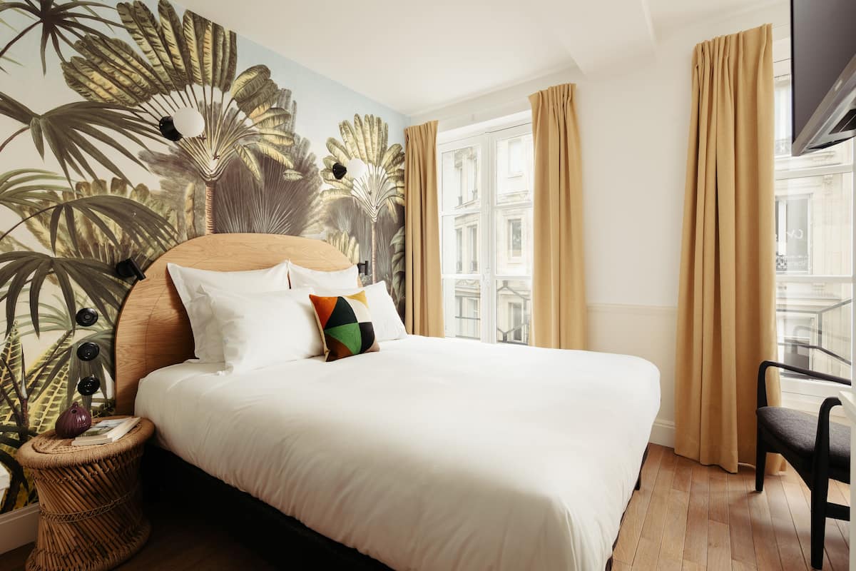 Ein Hotelzimmer mit einem großen Bett, Blick auf die Stadt durch das Fenster und einem Wandgemälde mit tropischen Motiven.