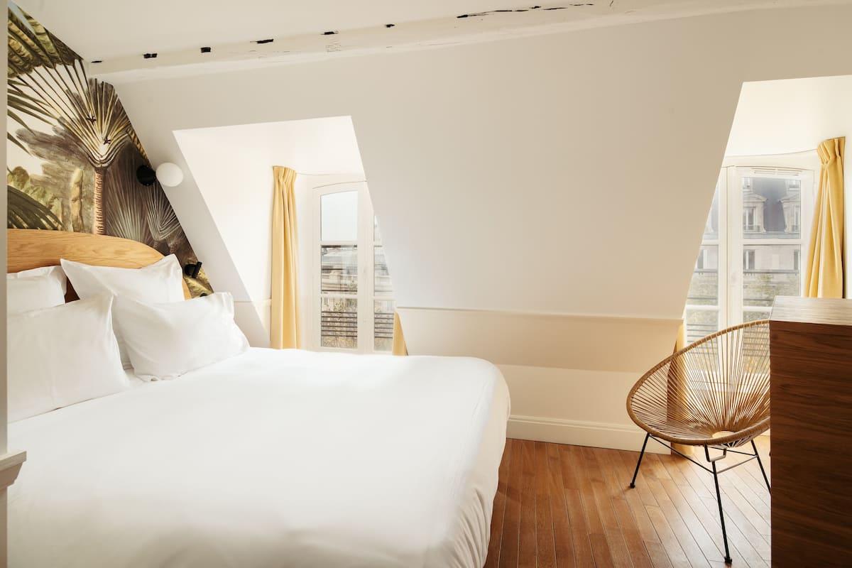 Ein Hotelzimmer mit einem großen Bett, einem Holz-Kopfteil, einem Rattan-Sessel und Blick auf die Stadt durch große Fenster.
