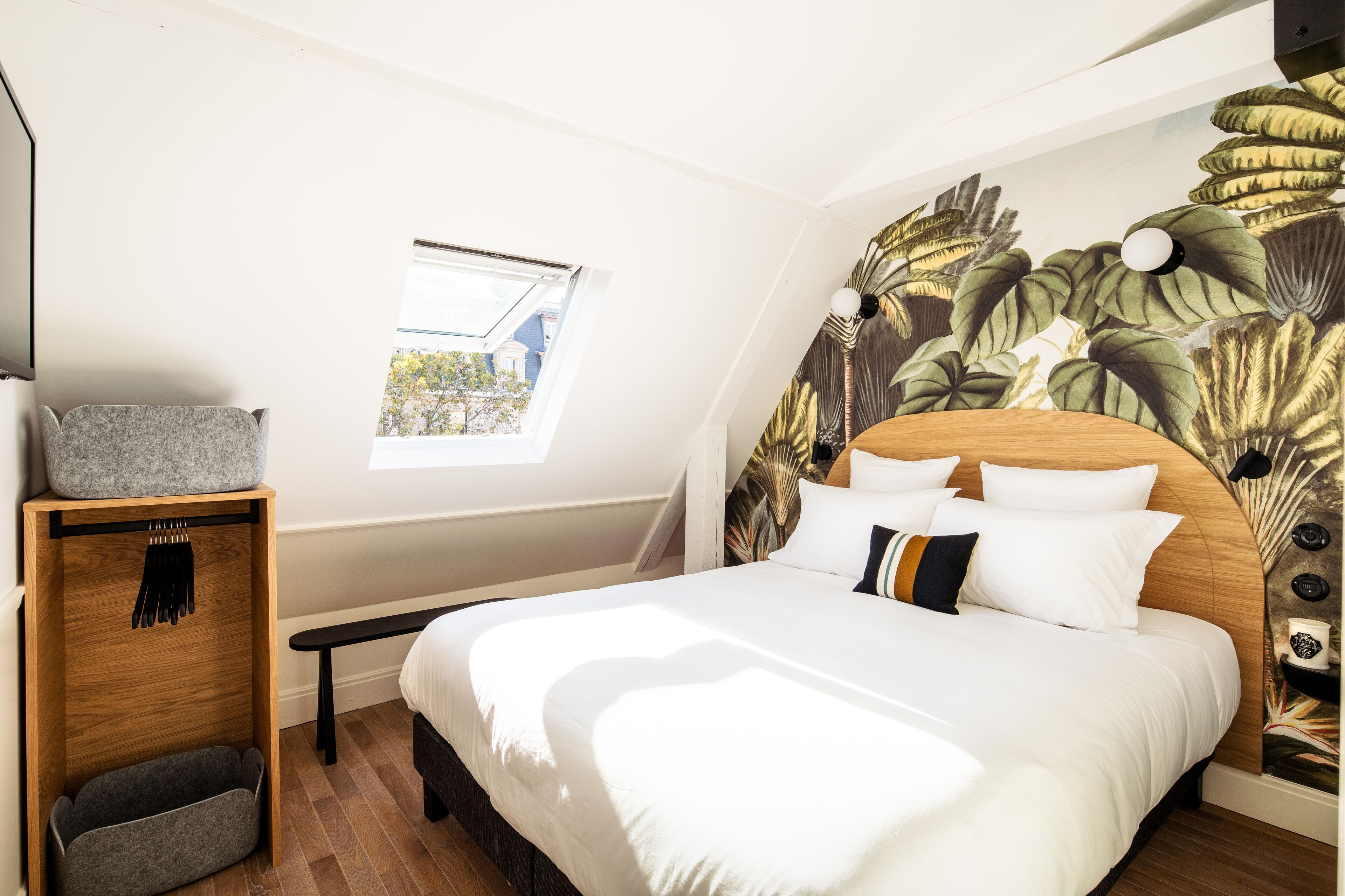 A bedroom with a large bed, a wooden headboard, a small window, and a wall with a tropical leaf mural.
