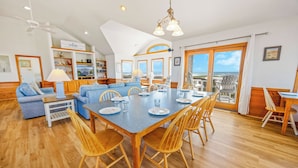 A spacious living area with a dining table, blue sofa, wooden chairs, and a view of the ocean through large windows. - Oceanfront Dog Friendly Home in Avon w/ Elevator, Hot Tub, Game Room, Wet Bar (Avon)