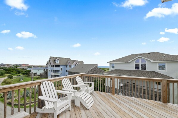 Une terrasse en bois, agrémentée de chaises Adirondack blanches, qui offre une vue sur un quartier résidentiel avec ses maisons et un ciel dégagé.