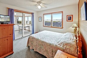 A bedroom with a bed, a wooden dresser, a television, a ceiling fan, a window with a view, and a door leading to a balcony.