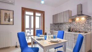 A modern kitchen with a gas stove, wooden cabinets, and a patterned backsplash. There is a dining table with blue chairs and a window with a view of the outside. - Cozy Syracuse House (Siracusa)