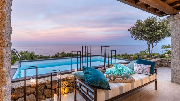 A poolside seating area with a view of the sea, stone walls, and a wooden pergola.