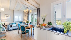 A bright kitchen with a large window, a dining area with a table and chairs, and a living room with a sofa and a chandelier.