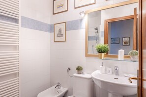 Un baño con azulejos blancos, un lavamanos blanco, un espejo, cuadros enmarcados y una pequeña planta en maceta.