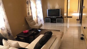 Una sala de estar con un sofá color beige, un plaid mullido negro, un adorno rosado, una silla, un televisor sobre un mueble bajo y una pequeña mesa.