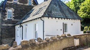 A small white building with a steeply pitched roof, a stone wall in front, and a larger stone building in the background. - Moorhurst Cottage (Grange-over-Sands)