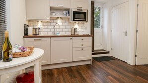 A modern kitchen with white cabinets, a microwave, and a sink. There is a wicker basket with items on a small table, and a red pot on the floor. - Moorhurst Cottage (Grange-over-Sands)