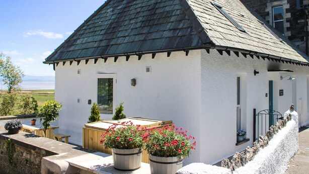 A small white building with a steeply pitched roof, a wooden deck, and potted plants. - Moorhurst Cottage (Grange-over-Sands)