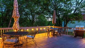 Una terraza de madera con una mesa, sillas, una parrilla y luces colgantes.