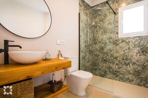 Un bagno con un mobile vanitario in legno, uno specchio rotondo, un lavabo bianco, un WC e una doccia con decori floreali.