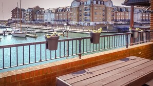 A waterfront balcony with a wooden bench, potted plants, and a view of a marina with boats and multi-story buildings in the background.