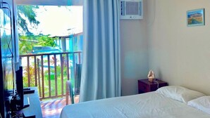A hotel room with a bed, a desk with a computer, an air conditioning unit, and a view of a balcony with a railing.