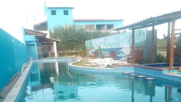 A swimming pool with a clear blue water surface, surrounded by a tiled deck and a covered area with lounge chairs.