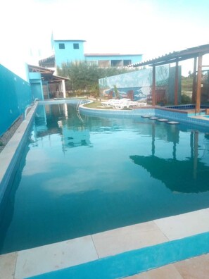 A swimming pool with a clear blue water surface, surrounded by a tiled deck and a covered area with lounge chairs.