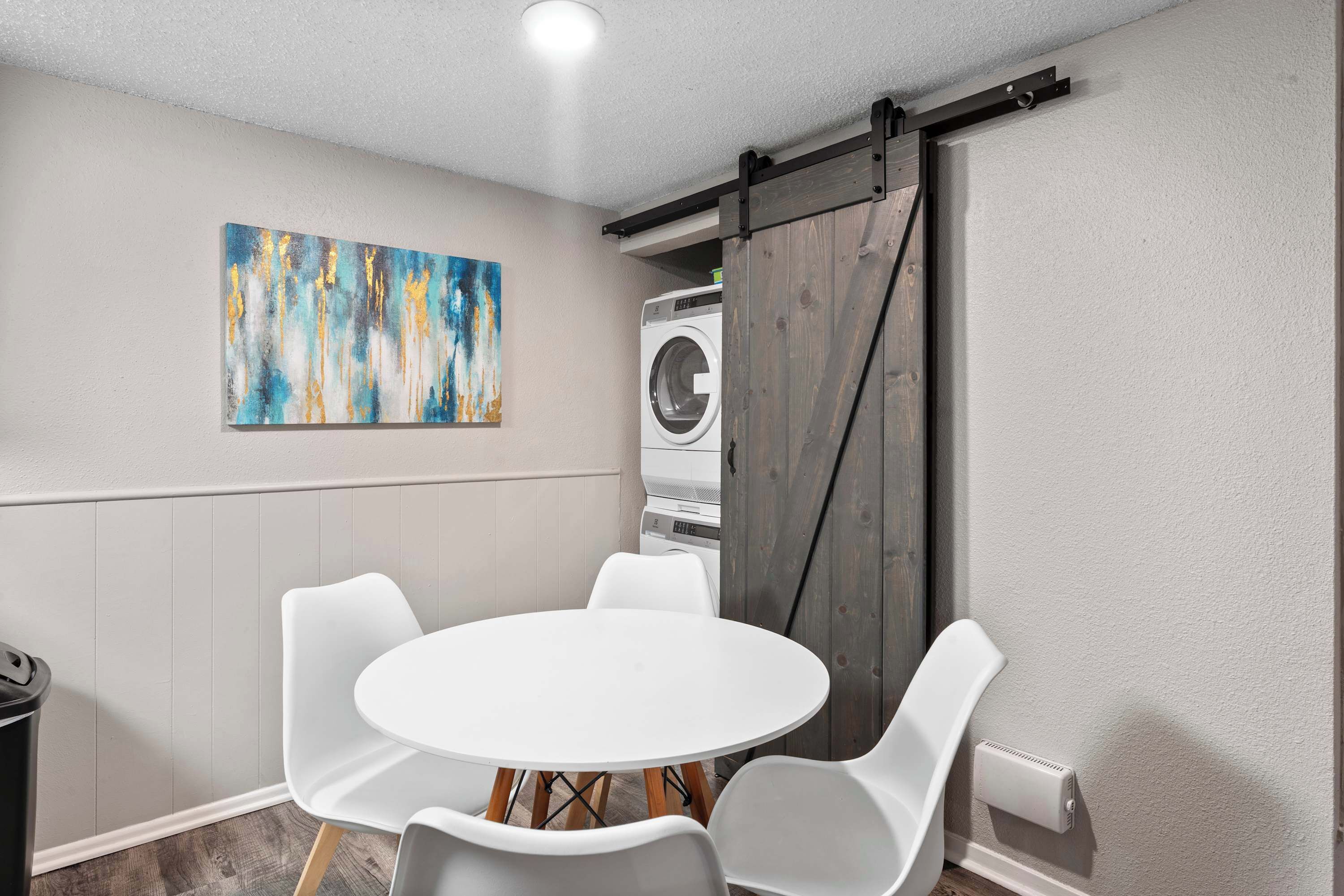 A room with a round white table, four white chairs, a large abstract painting, a washing machine, and a sliding barn-style door.